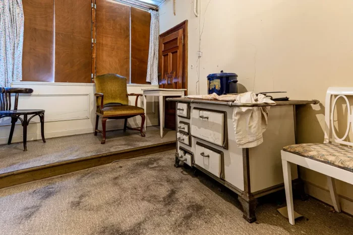 Ein altes, staubiges Zimmer mit gedämpfter Beleuchtung verfügt über eine Auswahl an Möbeln, darunter einen Holzstuhl, einen gepolsterten Sessel, einen Tisch und einen Vintage-Ofen. Die Fenster sind mit Holzbrettern bedeckt, und auf beiden Seiten sind lange Vorhänge.