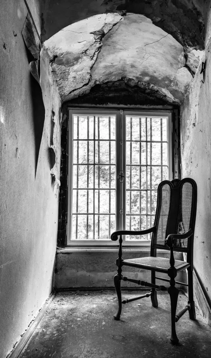 Schwarz-Weiß-Bild eines alten, verfallenen Raumes mit einem Gitterfenster und einem Vintage-Holzstuhl.