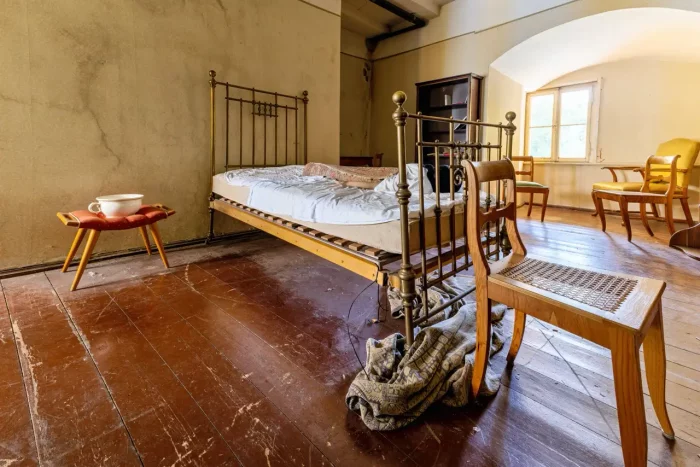 Ein altes, rustikales Schlafzimmer mit einem Messingbett, Holzboden, einem kleinen Tisch mit einer Schüssel und verstreuten Stühlen. Das Zimmer hat ein einziges Fenster mit natürlichem Licht.