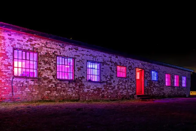 Ein rustikales Gebäude mit einer Backsteinfassade, die nachts von lila und rosa Lichtern beleuchtet wird. Die Struktur verfügt über mehrere Fenster und eine rote Tür mit einer grasbewachsenen Fläche davor.