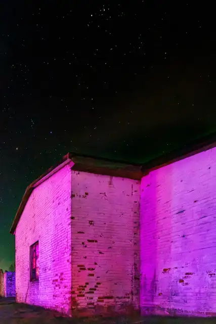 Ein hell beleuchtetes weißes Gebäude mit einem rosa Farbton steht vor einem Sternenhimmel. Das Bild erfasst die Ecke der Struktur und hebt ihre Textur und Farbkontraste mit dem dunklen Hintergrund hervor.