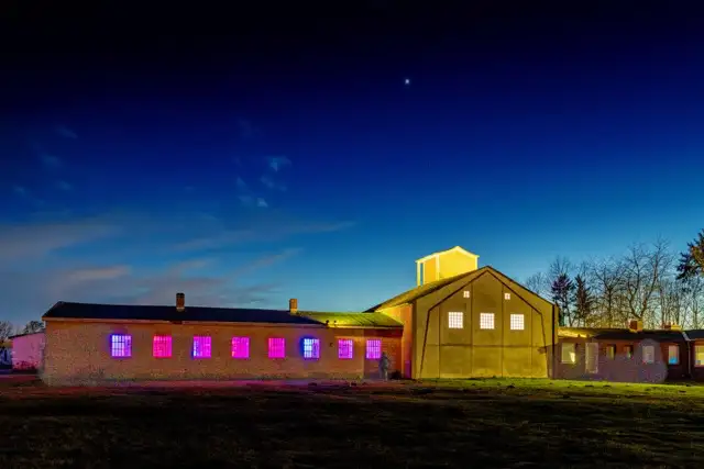Eine nächtliche Szene mit zwei Gebäuden mit hell beleuchteten Fenstern in Rosa- und Gelbtönen, die vor einem tiefblauen Himmel stehen. Sterne sind im Hintergrund zu sehen, was der Landschaft eine ruhige Atmosphäre verleiht.