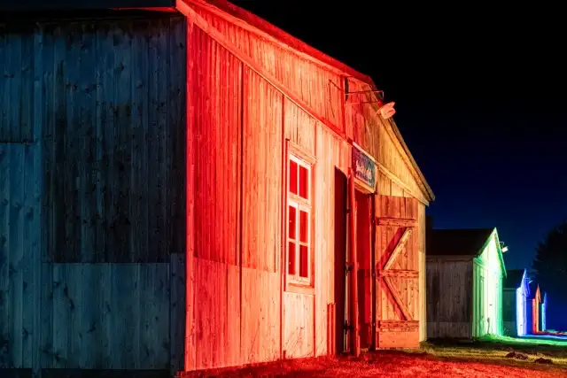 Ein Holzgebäude, das nachts mit roten und grünen Lichtern beleuchtet wird und kontrastierende Farben vor einem dunklen Himmel zeigt. Eine nahe gelegene Struktur ist im Hintergrund sichtbar.