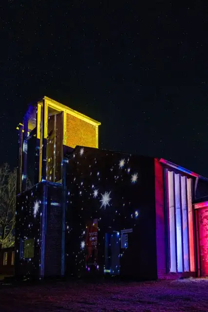 Ein Gebäude, das nachts mit bunten Projektionen von Sternen und Mustern an seiner Außenseite beleuchtet wird, vor einem dunklen Himmel voller entfernter Sterne.