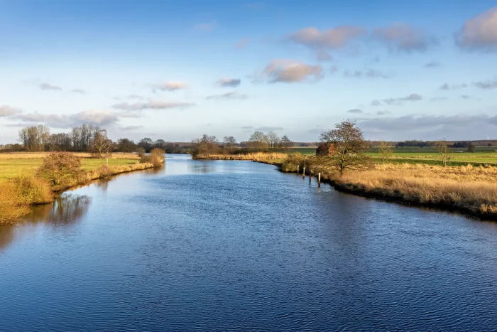 Eine ruhige Landschaft mit einem gewundenen Fluss, umgeben von grasbewachsenen Ufern und Bäumen. Der Himmel ist teilweise bewölkt, mit einem Hauch von Blau, der durchschaut. Ein kleines rotes Gebäude ist in der Ferne nahe dem Wasserrand zu sehen.