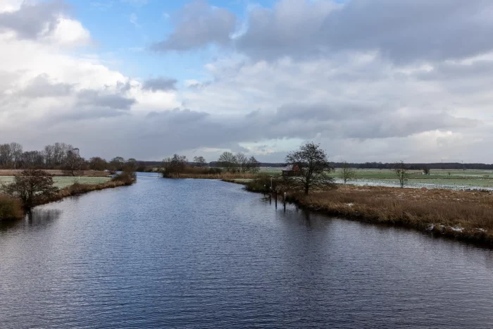 Eine ruhige Landschaft mit einem gewundenen Fluss, umgeben von grasbewachsenen Ufern und Bäumen. Der Himmel ist teilweise bewölkt, mit einem Hauch von Blau, der durchschaut. Ein kleines rotes Gebäude ist in der Ferne nahe dem Wasserrand zu sehen.