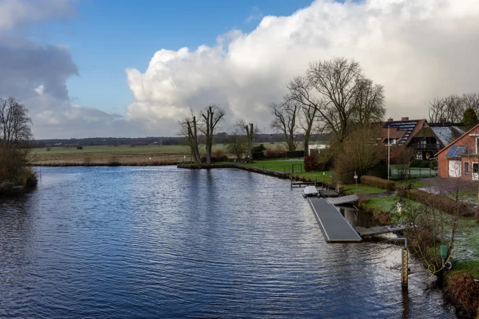 Eine ruhige Landschaft mit einem Fluss, der von grasbewachsenen Ufern, Bäumen und nahe gelegenen Häusern gesäuert wird. Der Himmel ist teilweise bewölkt mit hellblauen Flecken. Ein Holzdock erstreckt sich ins Wasser und verbessert die ruhige Umgebung.