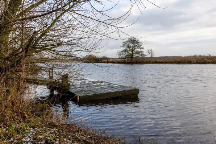 Ein Holzsteg erstreckt sich in einen ruhigen Fluss, umgeben von kahlen Bäumen und Gräsern unter einem bewölkten Himmel.