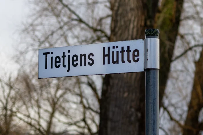 Ein Straßenschild mit der Aufschrift "Tietjens Hütte" ist an einer Stange angebracht, mit kahlen Bäumen im Hintergrund.