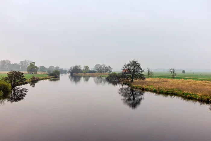 Eine ruhige Landschaft mit einem gewundenen Fluss, umgeben von grasbewachsenen Ufern und Bäumen. Der Himmel ist teilweise bewölkt, mit einem Hauch von Blau, der durchschaut. Ein kleines rotes Gebäude ist in der Ferne nahe dem Wasserrand zu sehen.