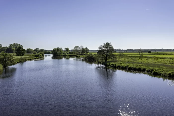 Eine ruhige Landschaft mit einem gewundenen Fluss, umgeben von grasbewachsenen Ufern und Bäumen. Der Himmel ist teilweise bewölkt, mit einem Hauch von Blau, der durchschaut. Ein kleines rotes Gebäude ist in der Ferne nahe dem Wasserrand zu sehen.