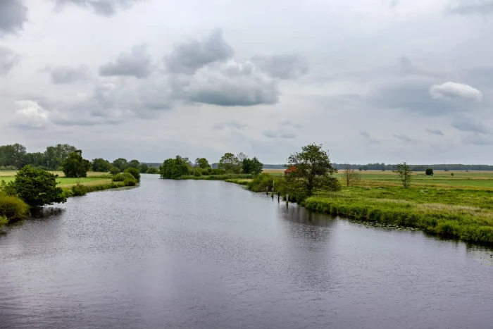 Eine ruhige Landschaft mit einem gewundenen Fluss, umgeben von grasbewachsenen Ufern und Bäumen. Der Himmel ist teilweise bewölkt, mit einem Hauch von Blau, der durchschaut. Ein kleines rotes Gebäude ist in der Ferne nahe dem Wasserrand zu sehen.