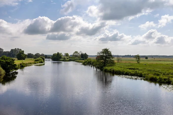 Eine ruhige Landschaft mit einem gewundenen Fluss, umgeben von grasbewachsenen Ufern und Bäumen. Der Himmel ist teilweise bewölkt, mit einem Hauch von Blau, der durchschaut. Ein kleines rotes Gebäude ist in der Ferne nahe dem Wasserrand zu sehen.