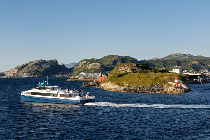 Fähre fährt an einer kleinen Insel mit Leuchtturm und Gebäuden vorbei, im Hintergrund felsige Hügel und klares Wetter