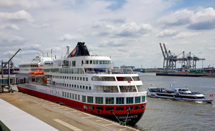Das Kreuzfahrtschiff Finnmarken der Hurtigruten liegt am Kai eines Hafens, im Hintergrund Containerkräne und ein kleineres Boot auf dem Wasser