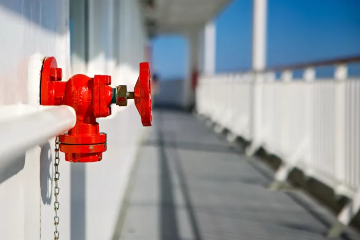 Rotes Ventil an der Außenwand eines Schiffsdecks mit weißem Geländer und blauem Himmel im Hintergrund
