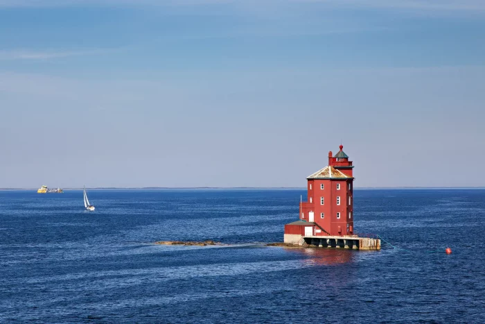 Roter Leuchtturm auf einer kleinen Insel im Meer unter klarem Himmel, Segelboot und Schiff in der Ferne
