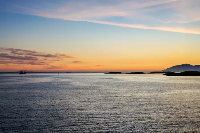 Fahrendes Hurtigruten-Schiff auf ruhiger See bei Sonnenuntergang mit Inseln und Bergen am Horizont