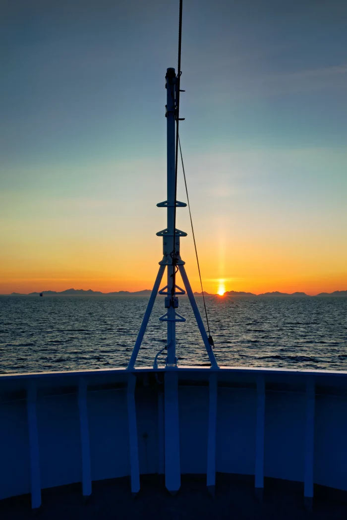 Sonnenuntergang über dem Meer vom Bug eines Schiffes mit Mast und Reling im Vordergrund