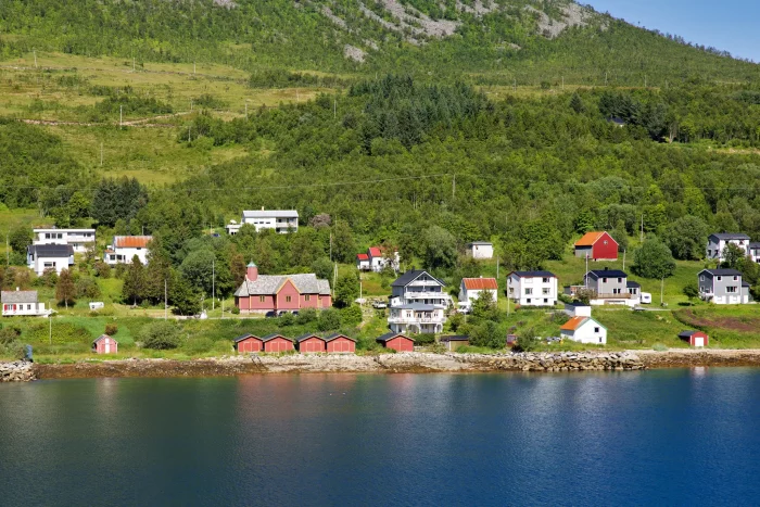 Kleine Siedlung mit bunten Häusern am Ufer eines ruhigen Gewässers, im Hintergrund bewaldeter Hang