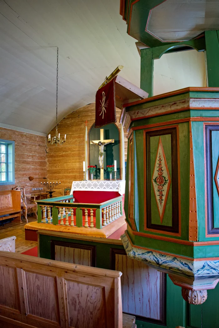 Innenraum einer kleinen Holzkirche mit bemalter Kanzel, Altar mit Kreuz und Kerzen, Holzbänken und Fenster mit Tageslicht
