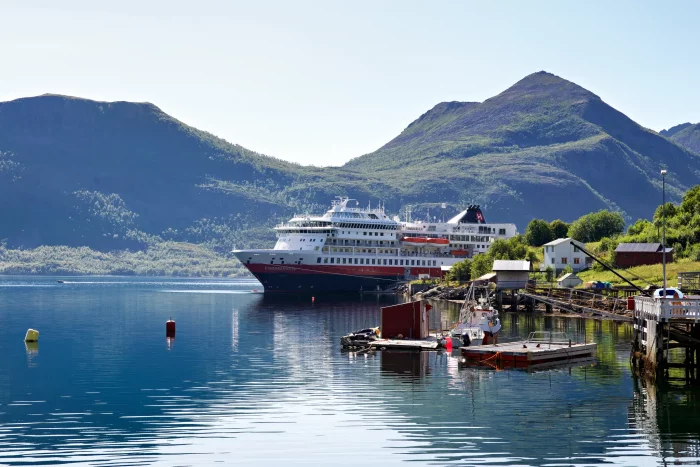 Hurtigruten-Kreuzfahrtschiff vor Fjord mit grünen Bergen und kleinem Hafen mit Booten