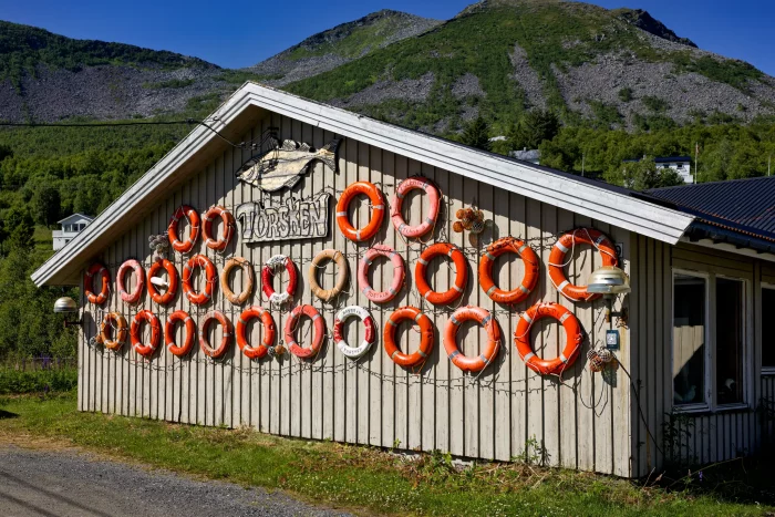 Holzhaus mit vielen orangefarbenen Rettungsringen an der Wand und einem Schild mit der Aufschrift 'TORSKEN' vor bewaldetem Berg