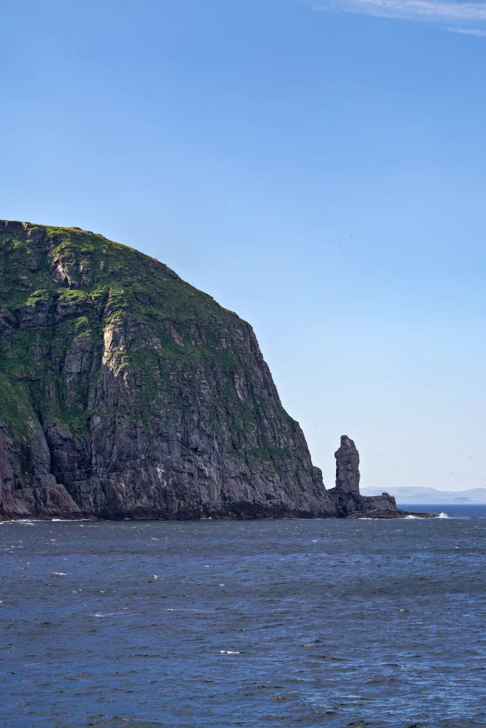 Steil aufragender Felsen mit grün bewachsenem Gipfel an der Küste, daneben ein markanter, schlanker Felsvorsprung im Meer