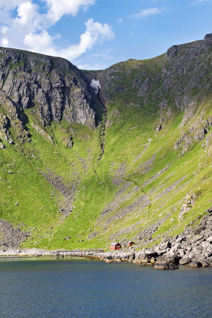 Kleine rote Holzhäuser am Ufer eines Fjords, umgeben von steilen, grünen und felsigen Bergen unter blauem Himmel