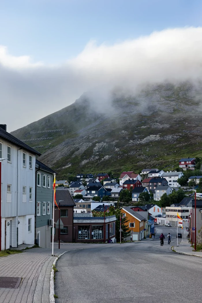 Straße mit bunten Häusern und zwei Radfahrern vor nebelverhülltem Berg in norwegischem Küstenort