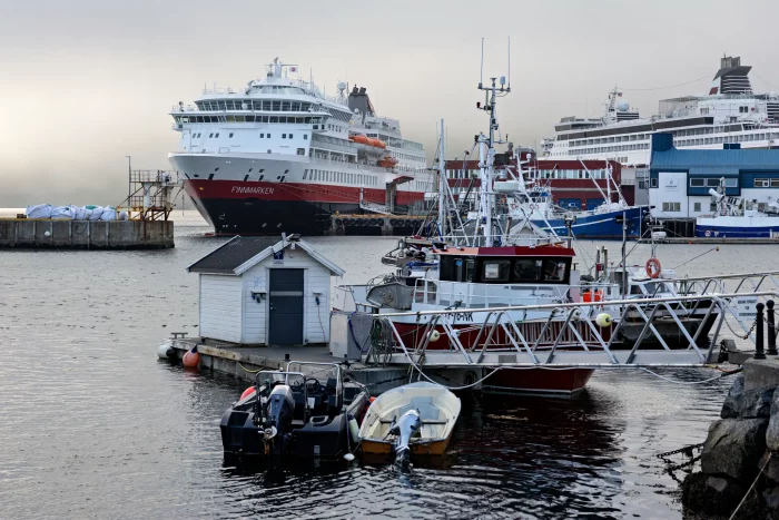 Hafen mit mehreren Schiffen, darunter das Hurtigruten-Kreuzfahrtschiff 'Finnmarken' im Hintergrund und kleinere Fischerboote im Vordergrund