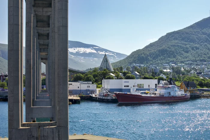 Rotes Hurtigruten-Schiff am Kai in einer norwegischen Küstenstadt mit Bergen und einer Kirche mit spitzem Dach im Hintergrund