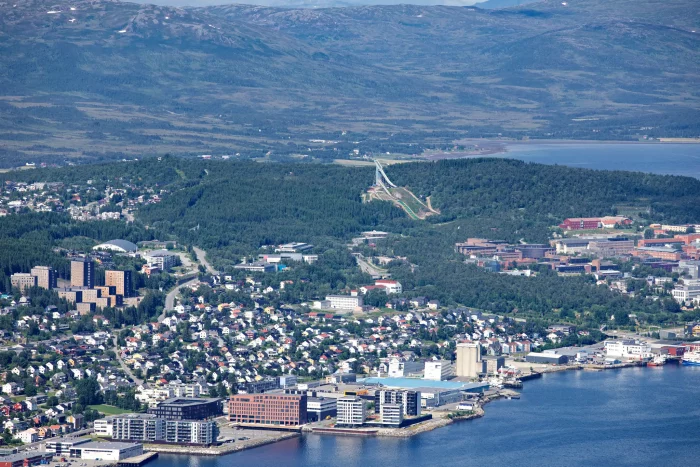 Luftaufnahme einer Küstenstadt mit Hafen, Wohn- und Geschäftsgebäuden, bewaldeten Hügeln und Bergen im Hintergrund