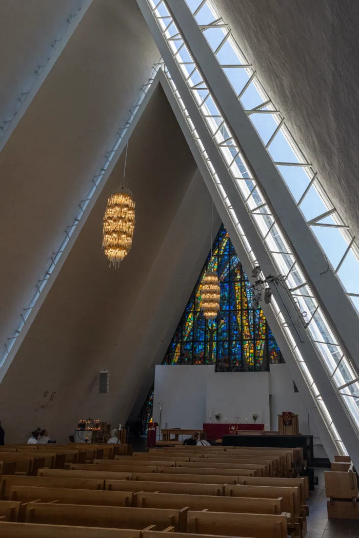 Innenraum der Eismeerkathedrale in Tromsø mit spitz zulaufender Decke, hängender Kronleuchter und buntem Glasfenster hinter dem Altar