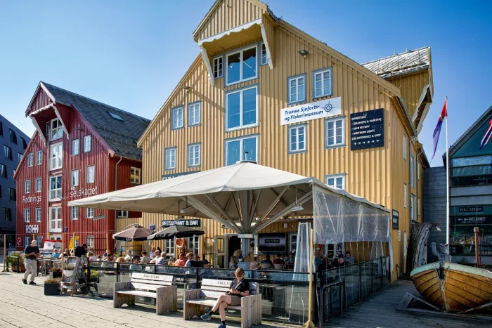 Gelbes historisches Gebäude mit Restaurantterrasse und Menschen, daneben rotes Gebäude am Hafen in Norwegen