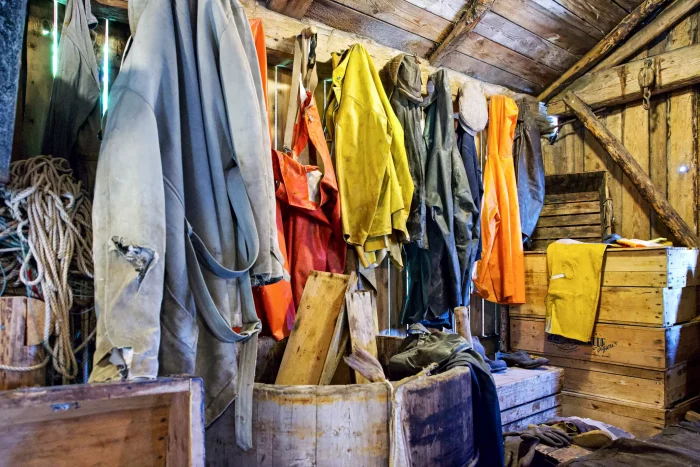 Mehrere wetterfeste Arbeitsjacken und Overalls hängen an einer Holzwand in einem rustikalen Raum mit Holzkisten und Seil