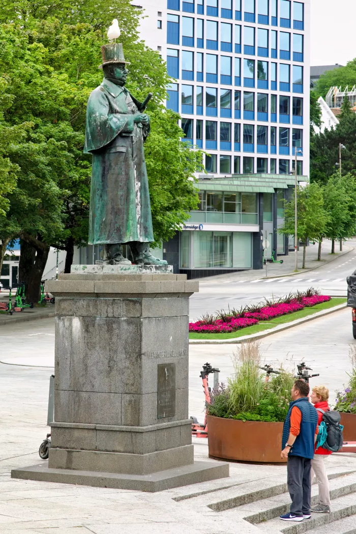Bronzestatue eines Mannes mit Zylinder und Mantel auf Steinsockel in urbanem Park, zwei Personen betrachten die Statue