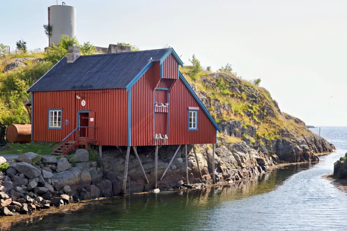Rotes Holzhaus auf Stelzen am felsigen Ufer eines ruhigen Gewässers unter klarem Himmel