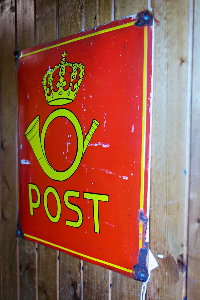 Rotes Metallschild mit gelber Posthorn-Ikone, Krone und dem Schriftzug 'POST' an einer Holzwand befestigt