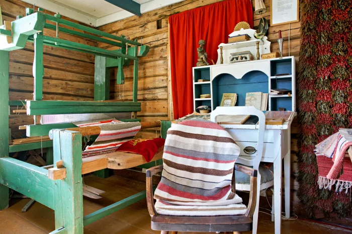 Traditionelle Webstühle und ein Holzstuhl mit gestreiftem Stoff in einem rustikalen Raum mit Holzwänden und roten Vorhängen