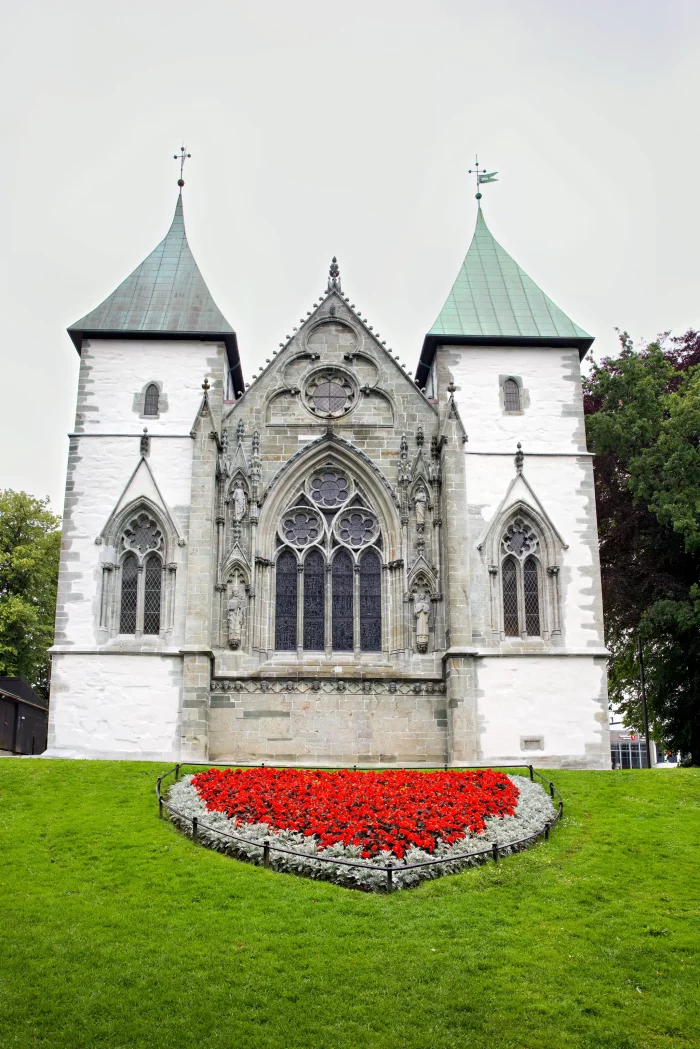 Gotische Kirche mit zwei Türmen und einem dreieckigen Giebel, davor ein Blumenbeet mit roten Blumen auf grünem Rasen