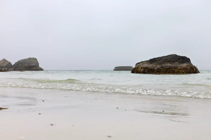Sandstrand mit mehreren großen Felsen im flachen Wasser unter grauem Himmel
