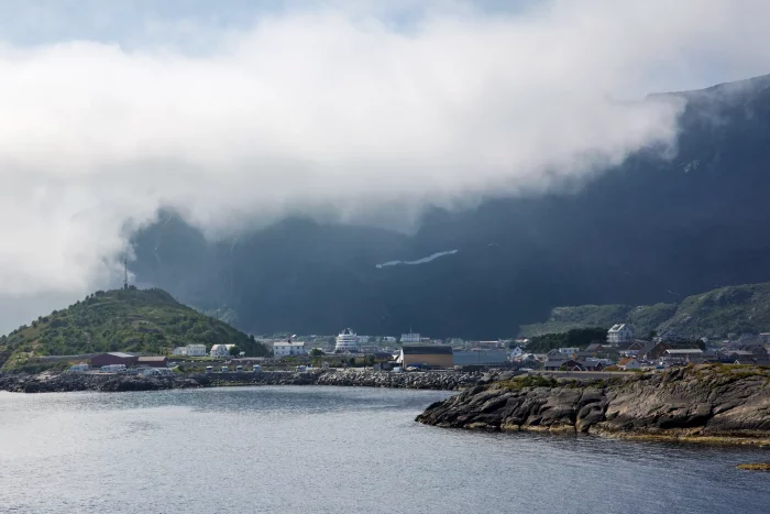 Küstenlandschaft mit kleinen Gebäuden, Felsen im Vordergrund und Bergen, die von Nebel bedeckt sind