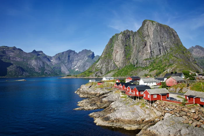 Rote Holzhäuser am felsigen Ufer eines Fjords mit hohen Bergen im Hintergrund unter blauem Himmel