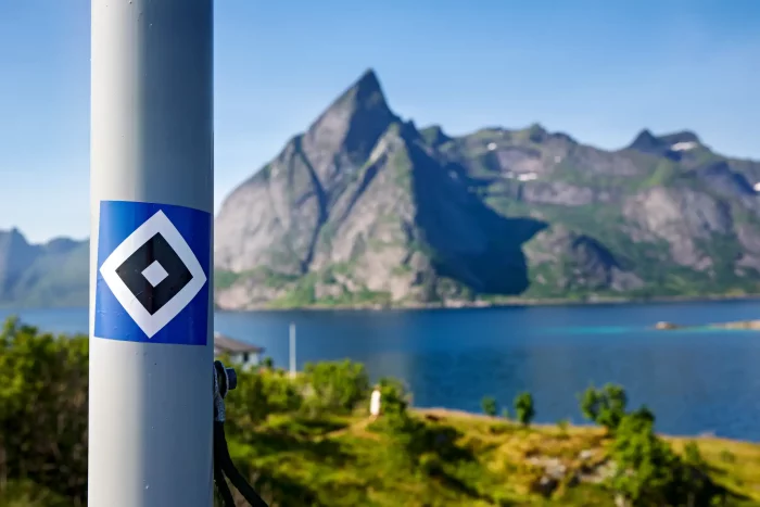 Blau-weiß-schwarze HSV-Raute vor Bergkulisse und Wasser