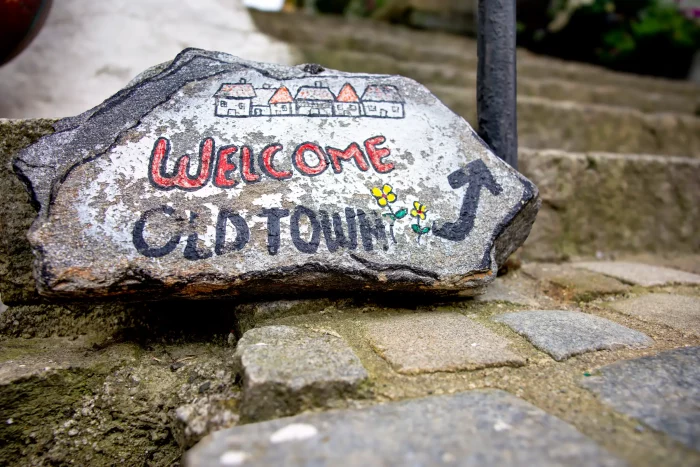 Stein mit handgemaltem Schriftzug 'Welcome Old Town' und Richtungspfeil, kleine Häuser und Blumen als Dekoration