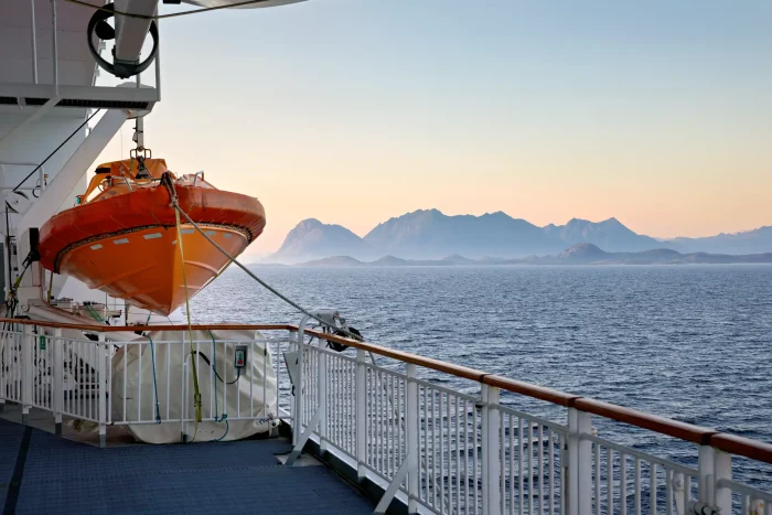 Blick von einem Schiff mit orangefarbenem Rettungsboot auf ruhiges Meer und Bergsilhouette bei Sonnenuntergang