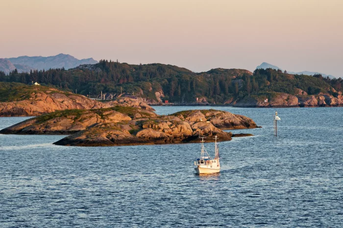 Kleines Fischerboot auf ruhigem Wasser vor felsigen Inseln mit bewaldeten Hügeln im Hintergrund bei Sonnenuntergang