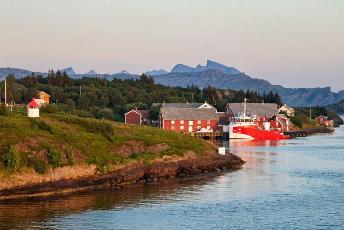 Rotes Schiff am Kai eines kleinen norwegischen Küstendorfes mit roten und weißen Häusern, bewaldeten Hügeln und Bergen im Hintergrund