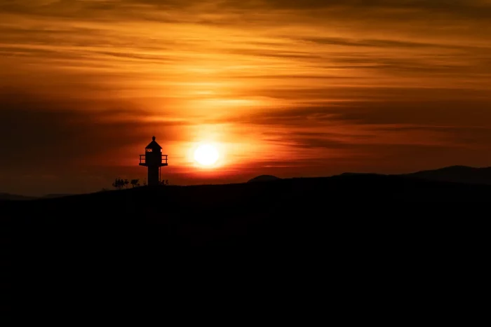 Leuchtturm auf Hügel vor orangefarbenem Sonnenuntergangshimmel